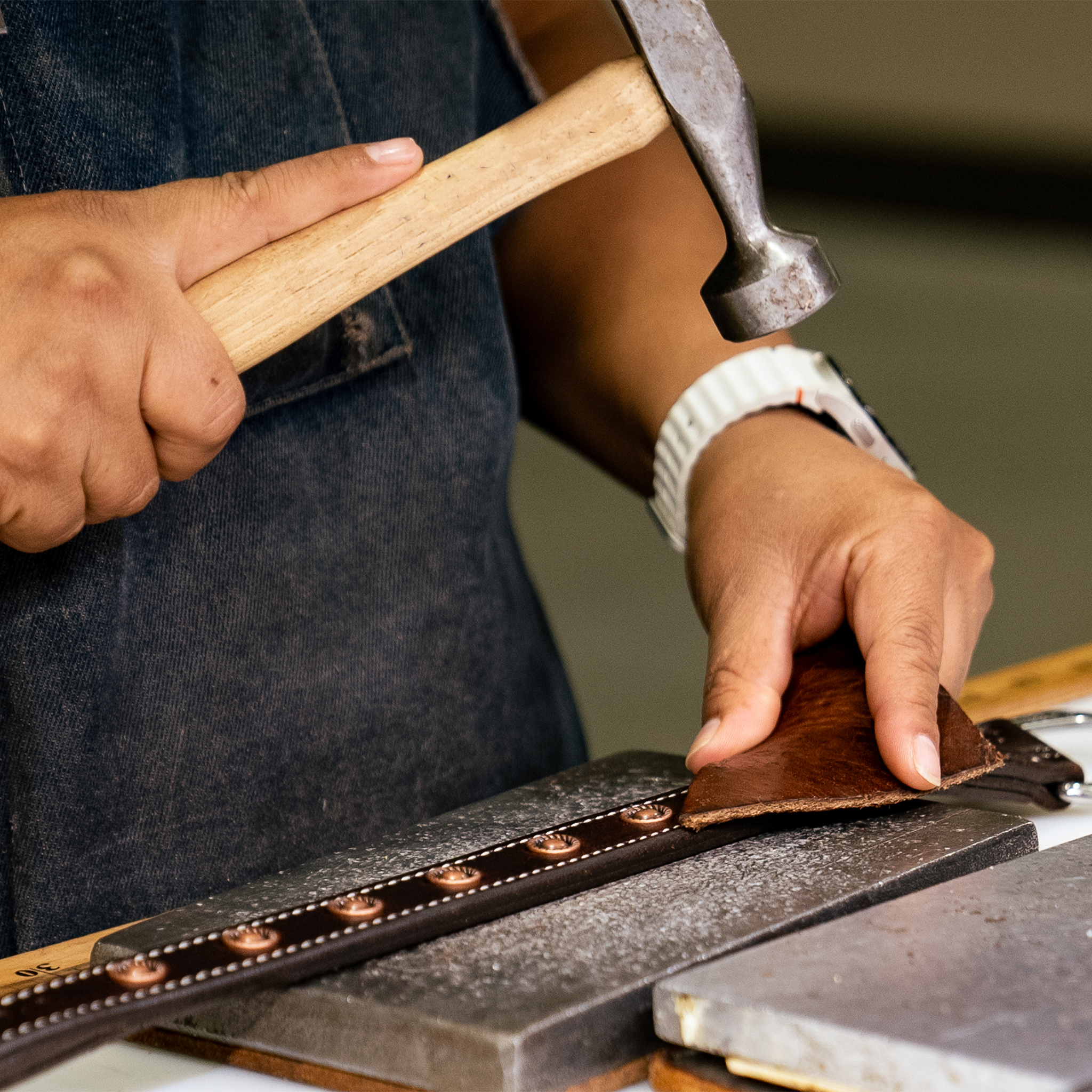 A craftsman in a denim apron hammers antique copper dots into a 1-inch chocolate leather breastcollar strap in the workshop. White stitching runs along both edges of the strap beside the row of rope edge dots.