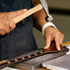 A craftsman in a denim apron hammers antique copper dots into a 1-inch chocolate leather breastcollar strap in the workshop. White stitching runs along both edges of the strap beside the row of rope edge dots.