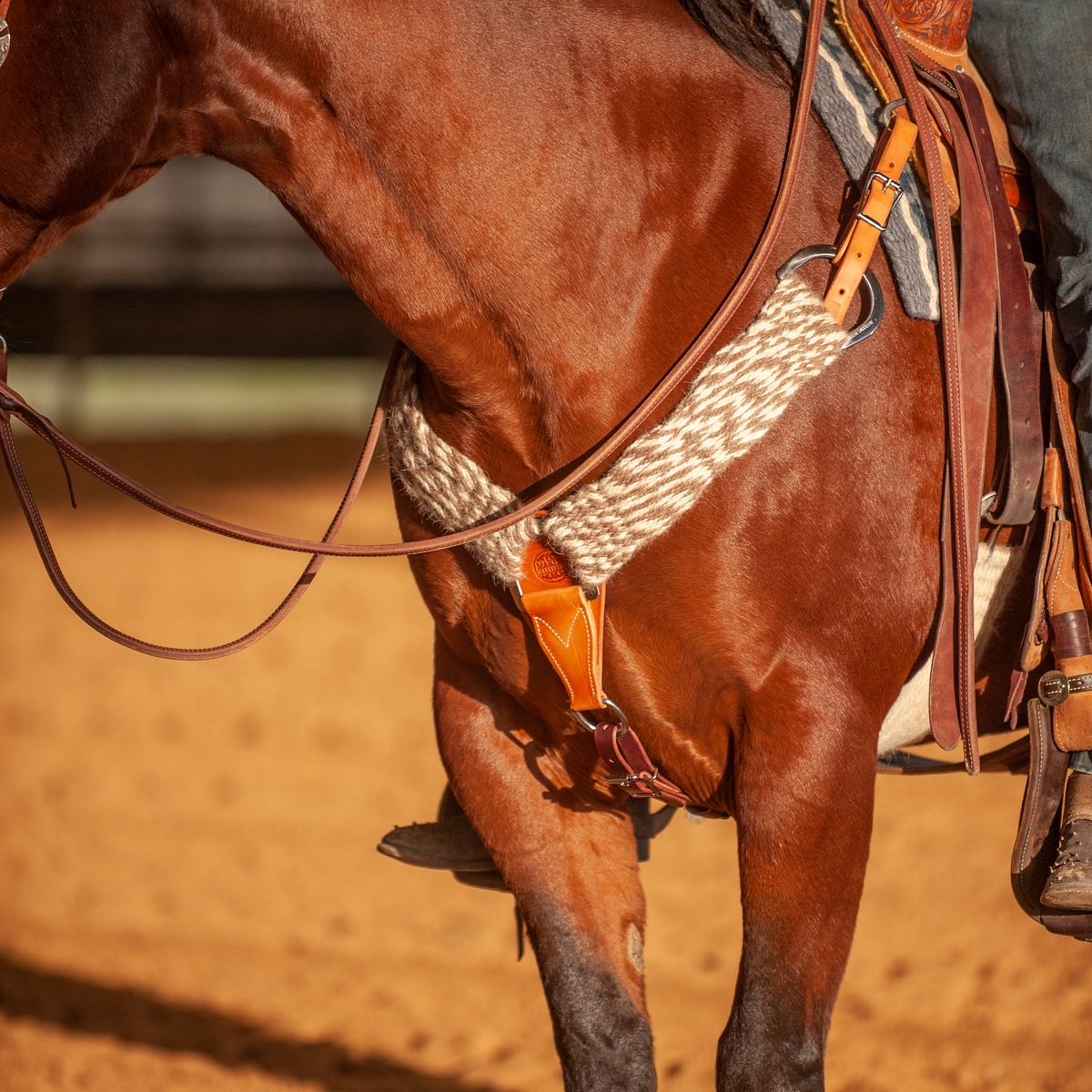 Natural mohair alpaca breastcollar on a bay horse in an arena, seen at the chest with a rider in the saddle. The 3-inch band of natural fiber strands runs to a leather center piece and up to a ring at the saddle.
