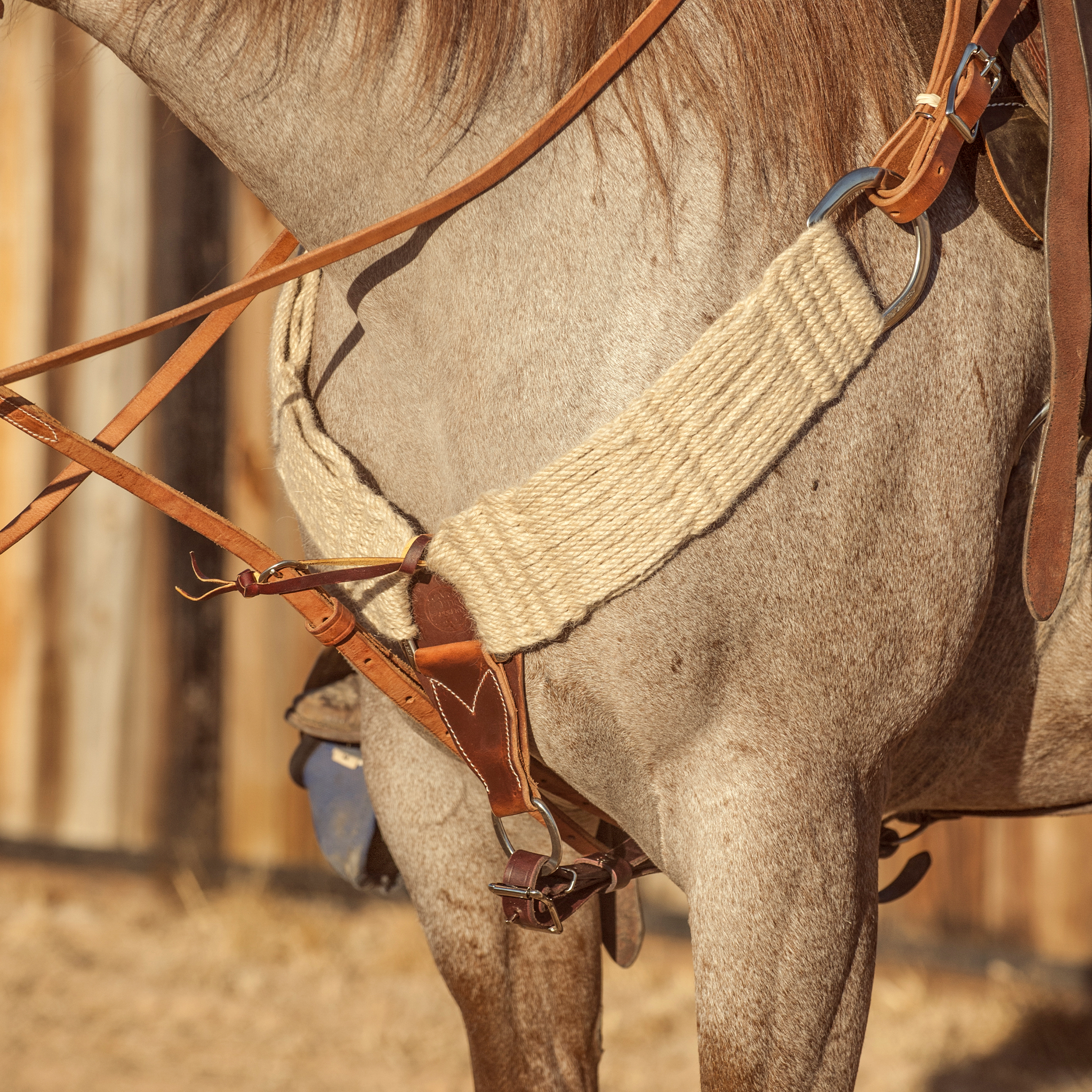 Natural mohair breastcollar on a roan horse standing by a wooden fence, seen at the chest. The 3-inch band of natural mohair strands runs from a ring at the saddle to a leather center piece with a tie-down strap.