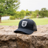 Black trucker cap with a white Martin Saddlery shield logo resting on a stone wall, with trees and a lawn behind it. The cap has a precurved bill and a black mesh back.
