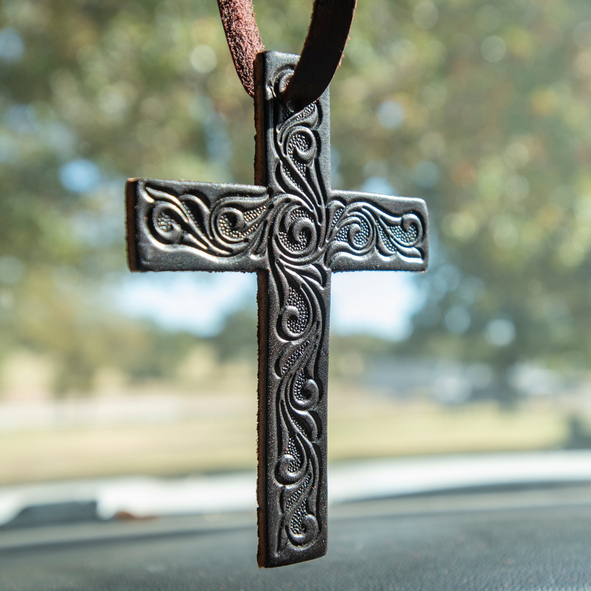 Black tooled leather cross air freshener hanging over a dashboard from a leather tie. Scroll tooling stands out across the arms and length of the cross.