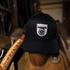 Black trucker cap with a white Martin Saddlery logo patch sitting on the horn of a tooled western saddle in a tack room. The cap is seen from the front.