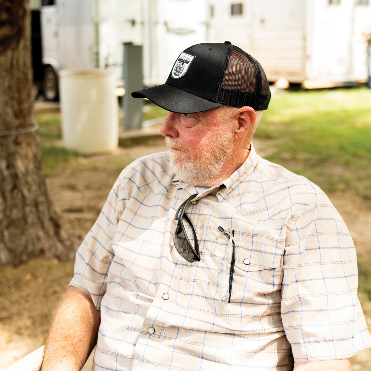 A man in a plaid shirt wearing a black trucker cap with a white Martin Saddlery logo patch, seen from the side outdoors. The mesh back and precurved bill of the cap are visible.