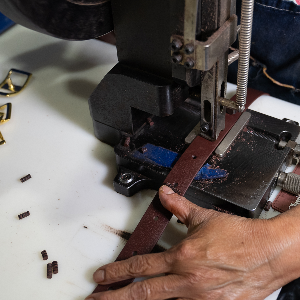 Hand guiding a brown BioThane tie-down strap through a punch press in the workshop. Punched pieces lie on the white bench beside the strap.
