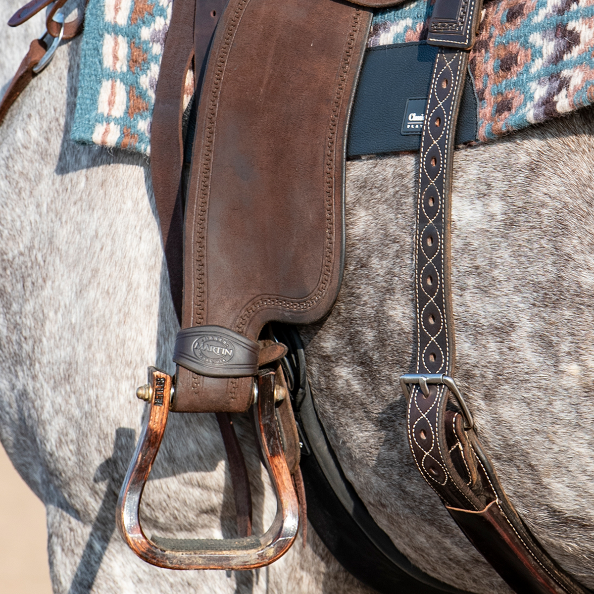 Chocolate stirrup hobble strap seen up close on a saddle fender above the stirrup, on a dapple gray horse. The strap wraps the stirrup leather and shows the Martin stamp.