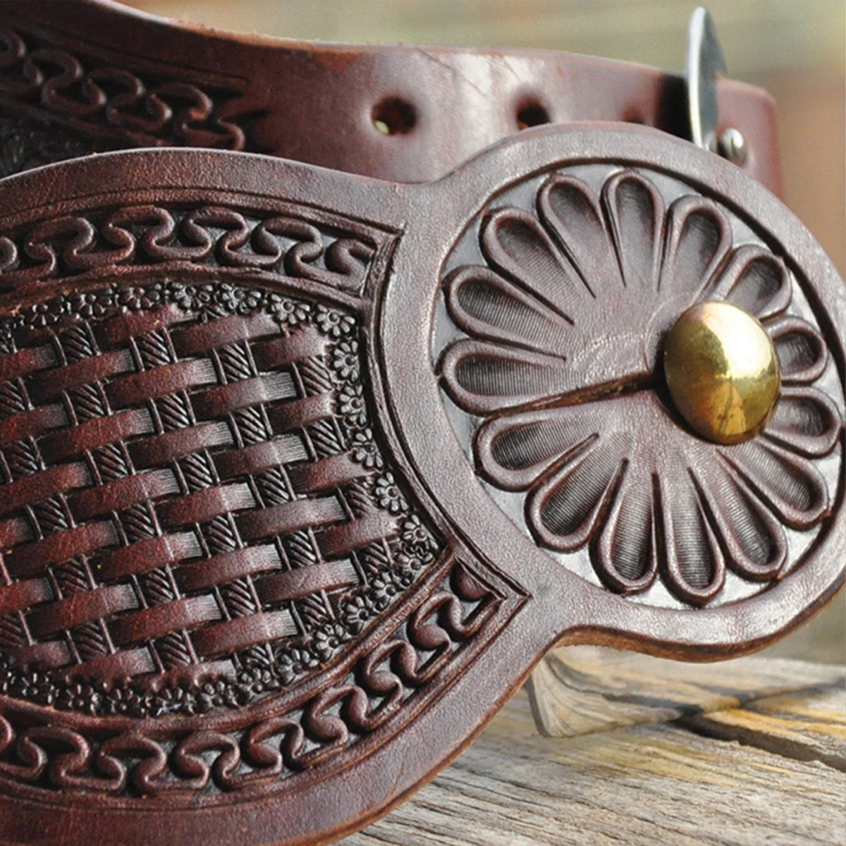 Chocolate Tombstone spur strap seen up close on a wood rail. A stamped flower surrounds the spur button beside the basket tooled center and San Carlos border.