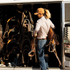 A woman in a clay performance cap lifts tack at an open trailer tack room filled with saddles. She wears the cap with a light shirt and jeans.