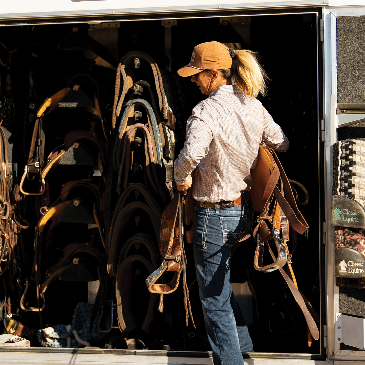 A woman in a clay performance cap lifts tack at an open trailer tack room filled with saddles. She wears the cap with a light shirt and jeans.