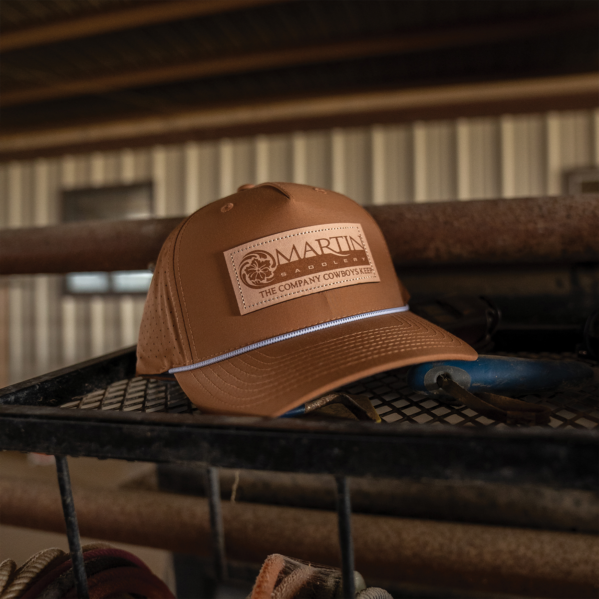 Clay performance hat resting on a wire rack in a barn. The leather patch logo reads Martin Saddlery, The Company Cowboys Keep, above a white rope across the bill.