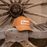 Clay rope hat with a leather Martin Saddlery patch sitting on a weathered wooden bench in front of a wagon wheel. A white rope runs across the bill.