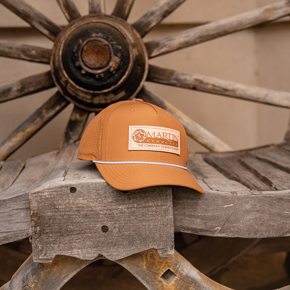 Clay rope hat with a leather Martin Saddlery patch sitting on a weathered wooden bench in front of a wagon wheel. A white rope runs across the bill.