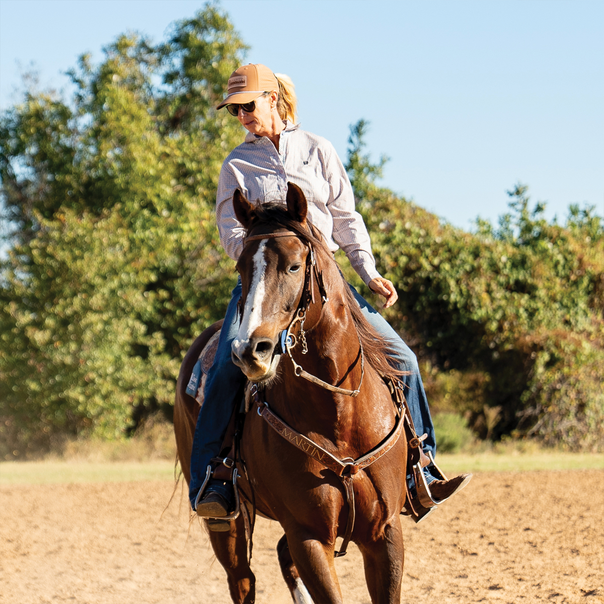 A rider in a clay cap and sunglasses sits on a sorrel horse in an outdoor arena, looking to one side. The western cap is worn with a ponytail pulled through the back.