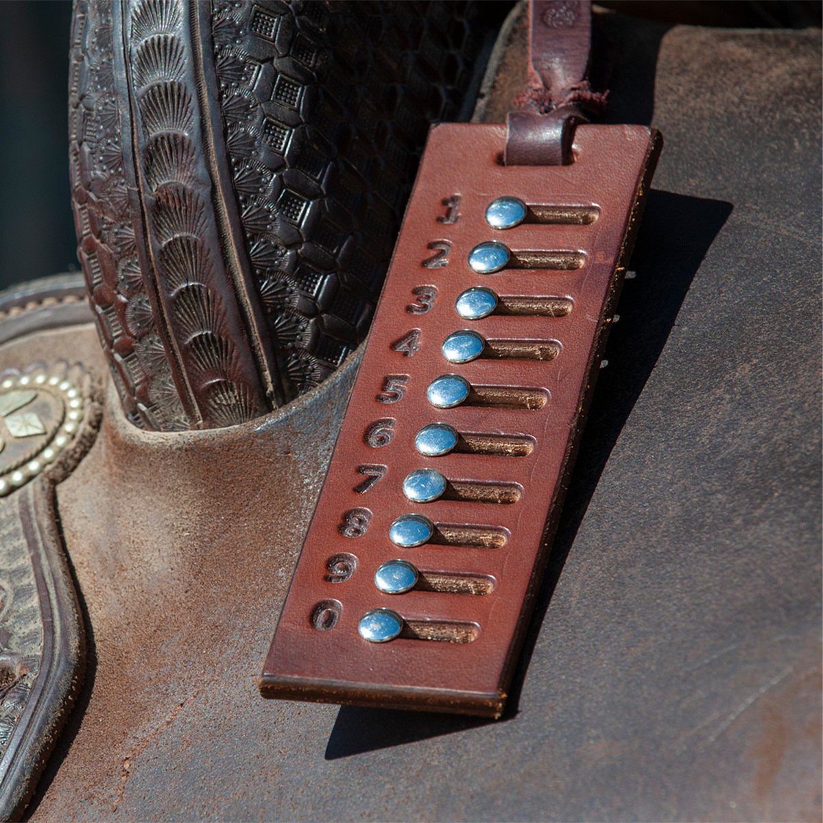 Leather cow counter resting on a saddle seat, hung from the saddle horn. Numbered slots hold metal rivets that slide to track which cows have been called in team penning and ranch sorting.