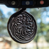 Round floral tooled leather air freshener hanging from a rearview mirror, with trees blurred behind. The dark leather shows a tooled flower inside swirling lines.