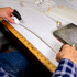 Craftsperson measuring white rope for a headsetter tiedown against a ruler on a workbench. Hands hold the rope beside a metal ring, with more cut rope lengths at the back of the bench.