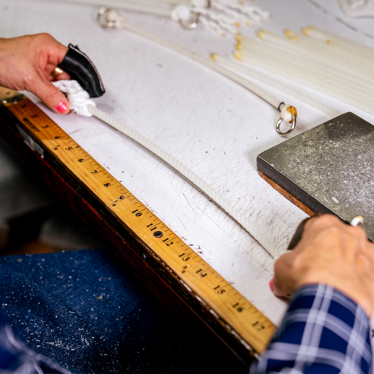 Craftsperson measuring white rope for a headsetter tiedown against a ruler on a workbench. Hands hold the rope beside a metal ring, with more cut rope lengths at the back of the bench.