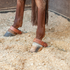 Kick chains on both hind legs of a sorrel horse standing in a bedded stall. Leather straps with rivets sit above the hooves, and the chains rest in the shavings.