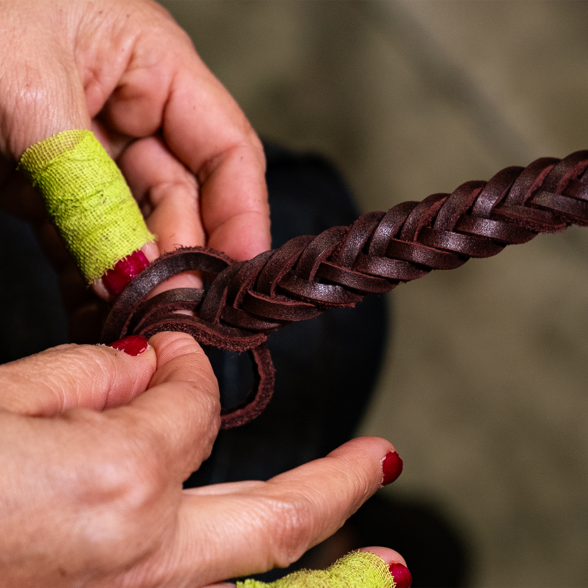 Craftsperson's hands plaiting chocolate latigo into a braided roping rein. One finger is wrapped in tape as the strands are worked into the 5 plait braid.