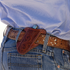 Floral tooled angled knife scabbard worn on a tooled leather belt at the hip of a person in jeans. A folding knife sits in the scabbard, which is edged in white stitching.