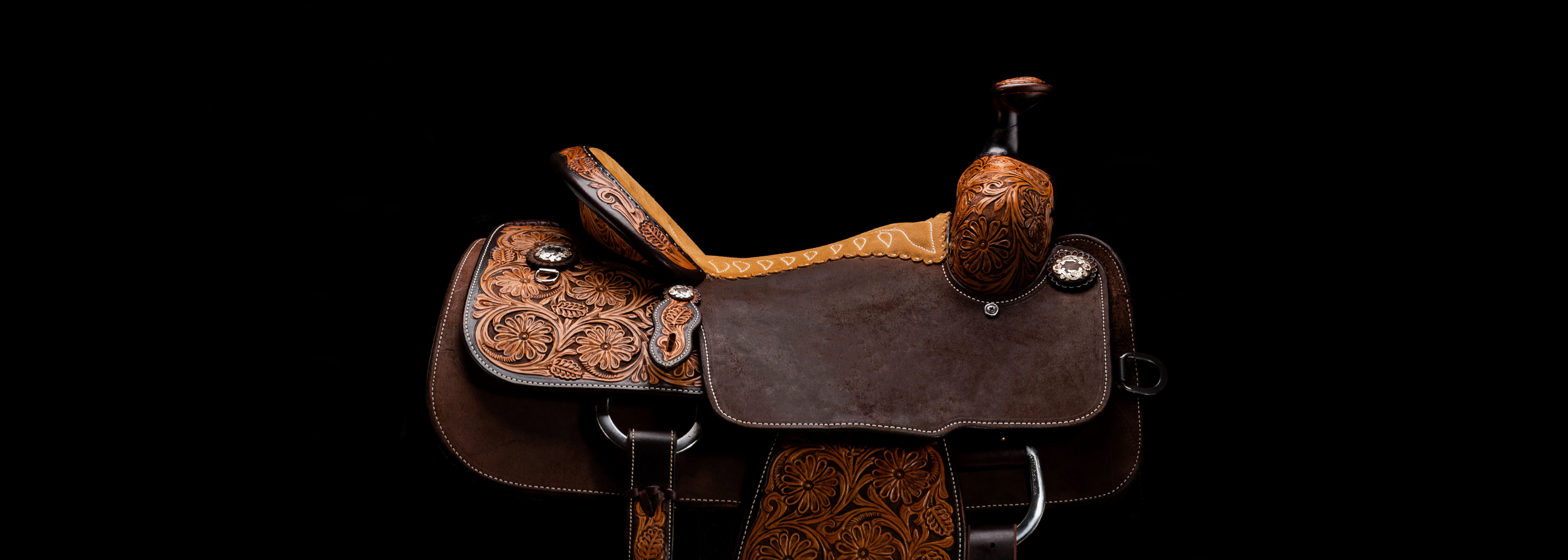 A detailed brown leather western saddle with intricate hand-tooled floral patterns and silver conchos, set against a solid black background.