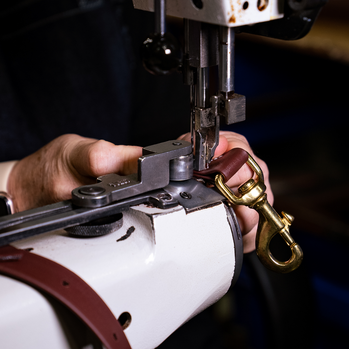 Hands stitching a snap onto a brown BioThane tie-down strap at a sewing machine in the workshop. The strap end folds through the snap under the needle.