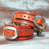 Chestnut square sliding spur straps stacked on painted boards, seen up close. A stamped border runs along each wide strap, and the sliding strap fastens with a stainless steel buckle.
