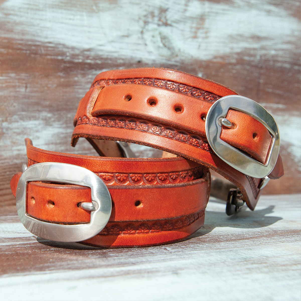 Chestnut square sliding spur straps stacked on painted boards, seen up close. A stamped border runs along each wide strap, and the sliding strap fastens with a stainless steel buckle.