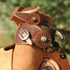 Chocolate buckaroo spur straps with basket tooling on a pair of spurs hung over a saddle horn. The tooled straps and their buckles show against a roughout saddle and green trees.