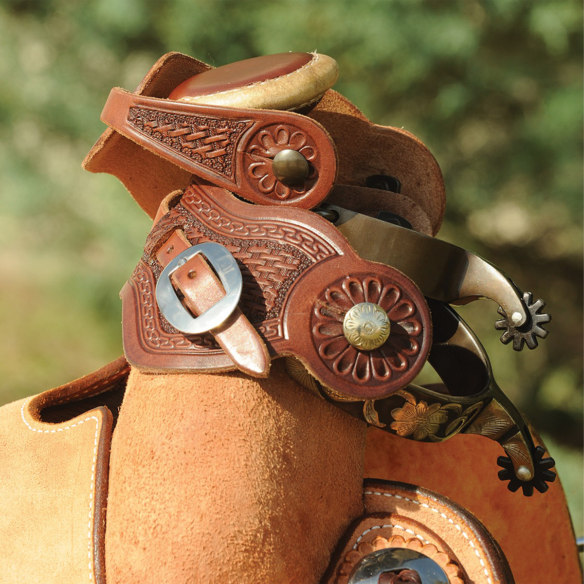Chocolate buckaroo spur straps with basket tooling on a pair of spurs hung over a saddle horn. The tooled straps and their buckles show against a roughout saddle and green trees.