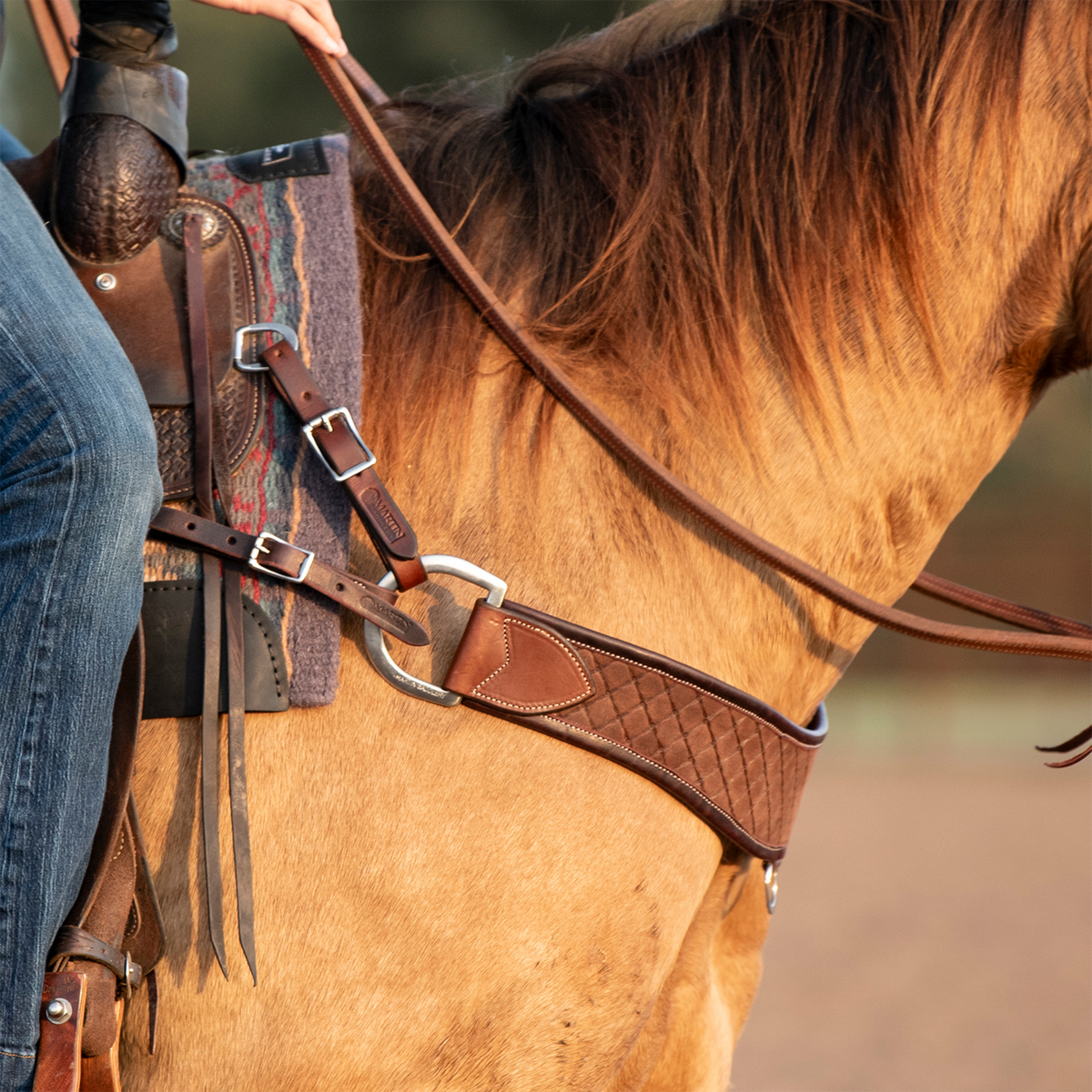 Martin Saddlery chocolate steer roper breastcollar on a buckskin horse, seen at the shoulder with a rider in the saddle. The quilted strap ends in a smooth leather tab at a ring, with buckled tug straps running to the saddle.