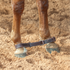 Double-stitched latigo hobbles from Martin Saddlery buckled around a horse's front pasterns in arena sand. White stitching runs along the dark leather straps.