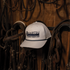 White Five Panel Rope Hat x Martin Saddlery hanging on a wooden tack room wall among leather bridles. The cap has a dark embroidered logo and a braided rope across the bill.