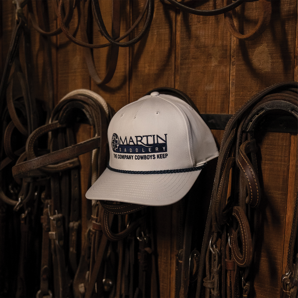 White Five Panel Rope Hat x Martin Saddlery hanging on a wooden tack room wall among leather bridles. The cap has a dark embroidered logo and a braided rope across the bill.