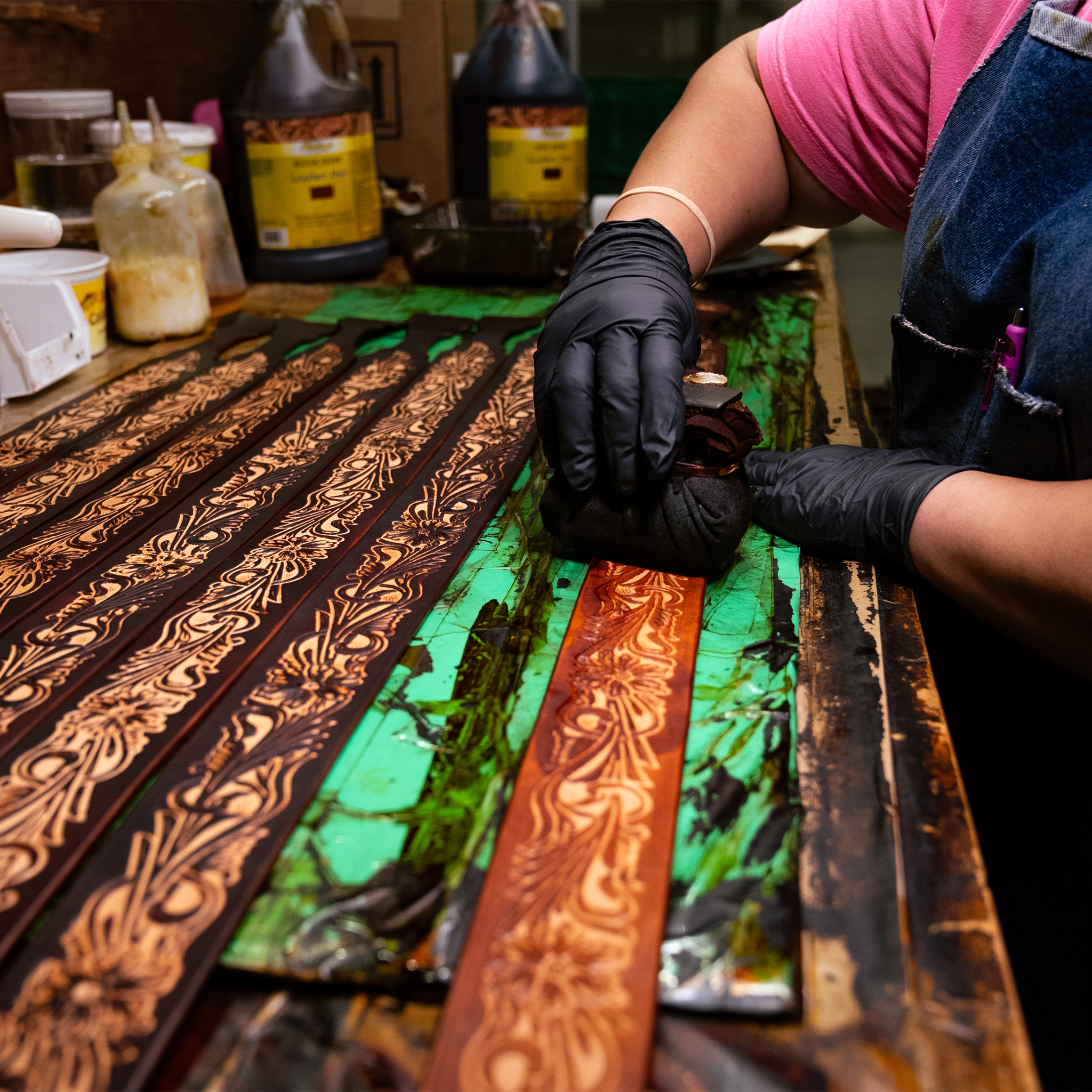 Craftsperson in black gloves dyeing floral tooled leather straps on a workbench in the workshop. Rows of tooled straps wait beside bottles of dye and finish.