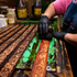 Craftsperson in black gloves dyeing floral tooled leather straps on a workbench in the workshop. Rows of tooled straps wait beside bottles of dye and finish.