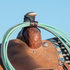 Latigo rope strap from Martin Saddlery holding a coiled green rope at the horn of a tooled saddle, against a blue sky. The dark leather strap wraps around the rope coils.
