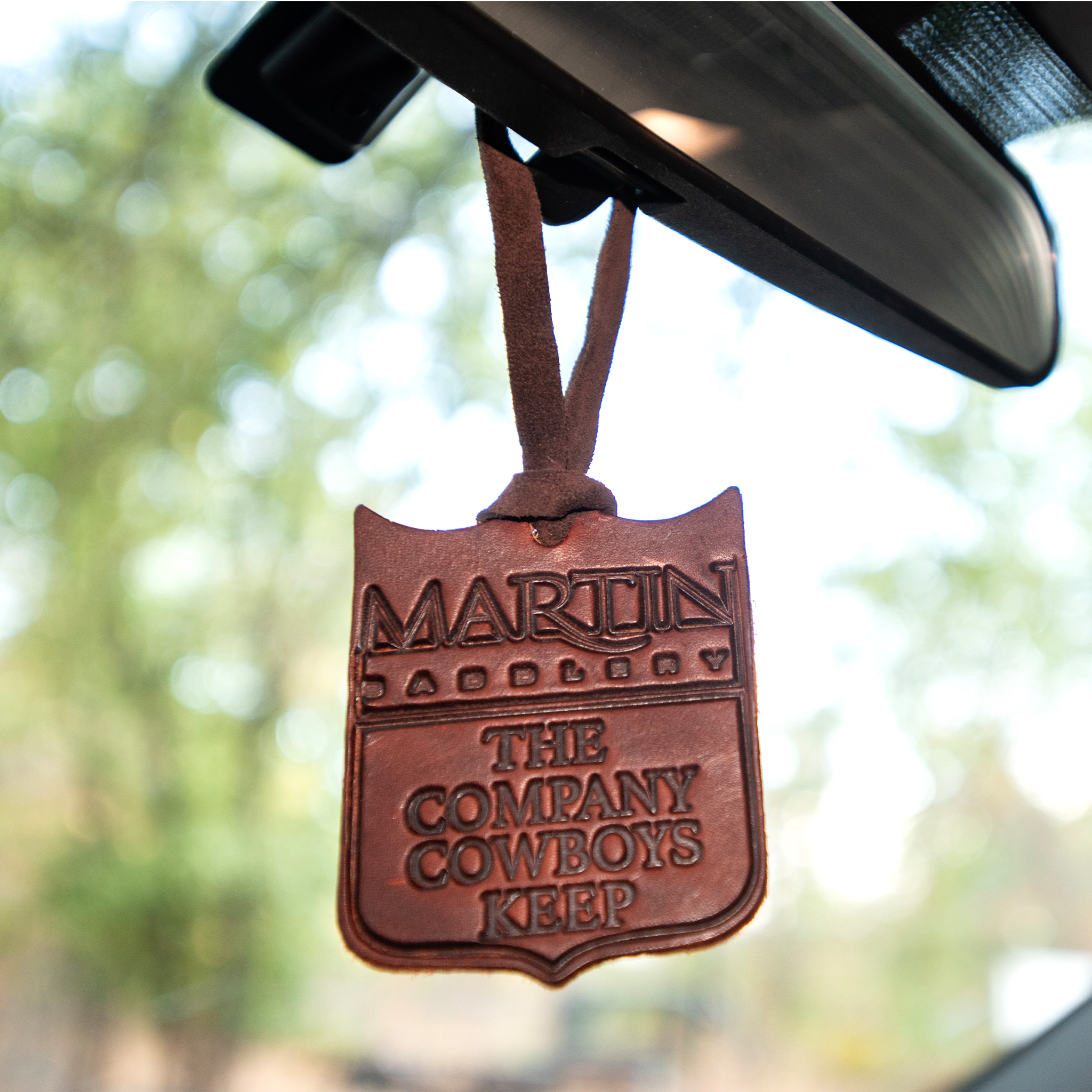 Martin Saddlery leather air freshener hanging from a rearview mirror, with trees blurred behind. The shield shaped leather is stamped Martin Saddlery, The Company Cowboys Keep.