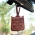 Martin Saddlery leather air freshener hanging from a rearview mirror, with trees blurred behind. The shield shaped leather is stamped Martin Saddlery, The Company Cowboys Keep.