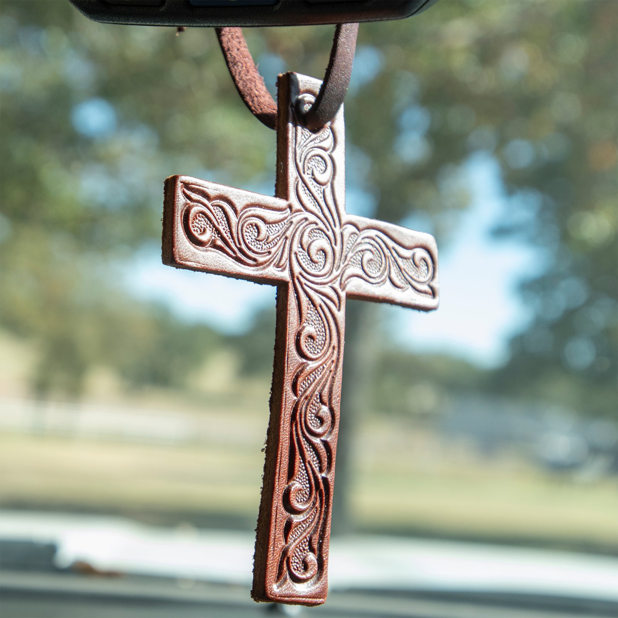 Tooled leather cross air freshener hanging from a rearview mirror, with trees and a field blurred behind. Scroll tooling covers the brown leather cross.