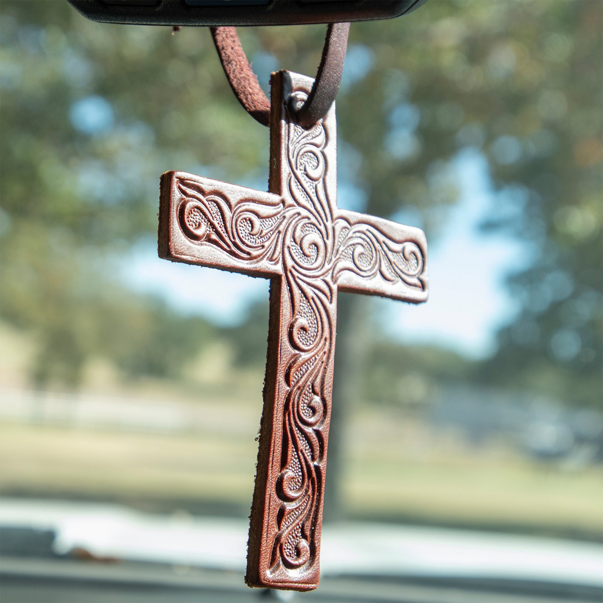 Tooled leather cross air freshener hanging from a rearview mirror, with trees and a field blurred behind. Scroll tooling covers the brown leather cross.