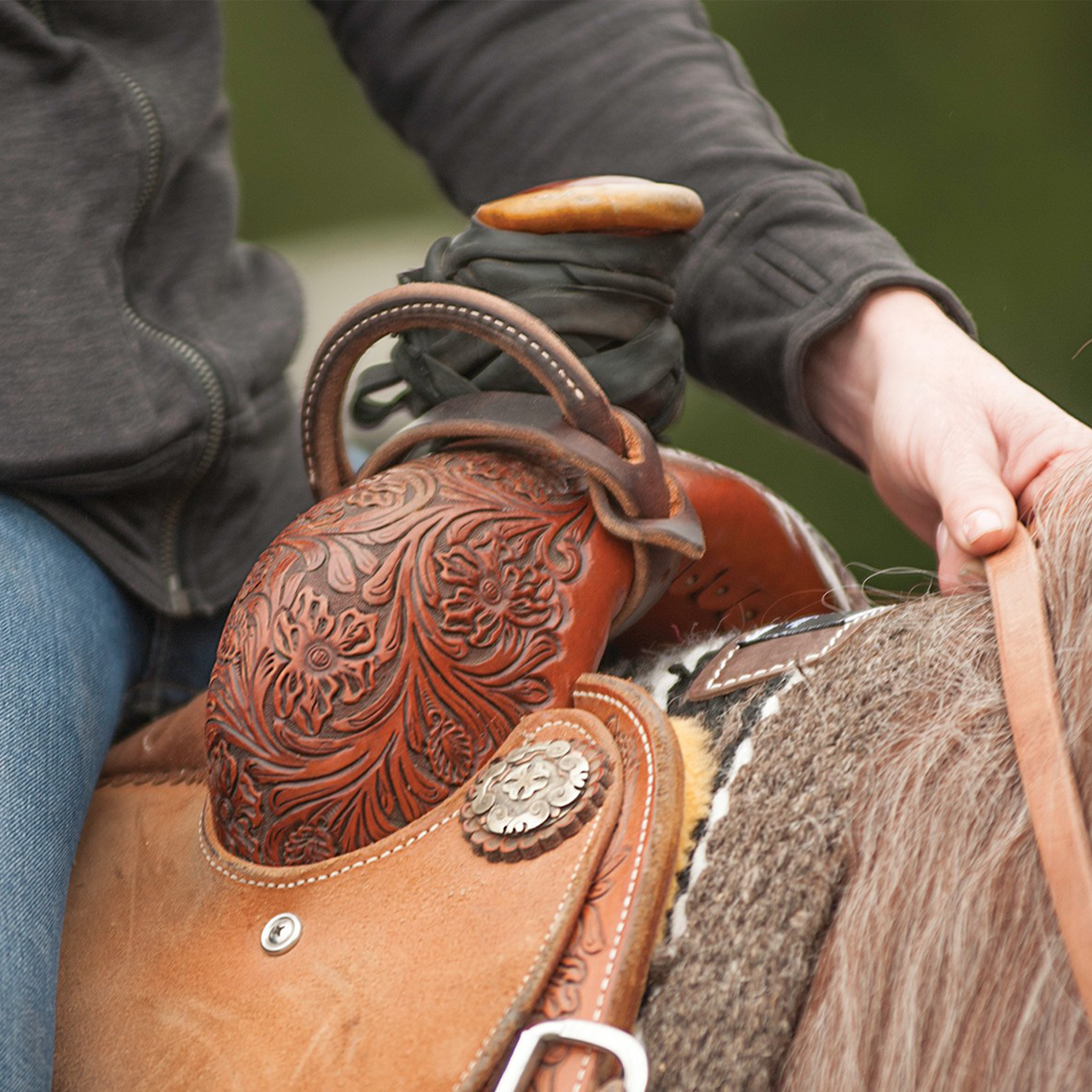 Leather night latch buckled around the swells of a floral tooled saddle, with a rider's hand resting beside it. The rolled leather handle sits in front of the saddle horn.