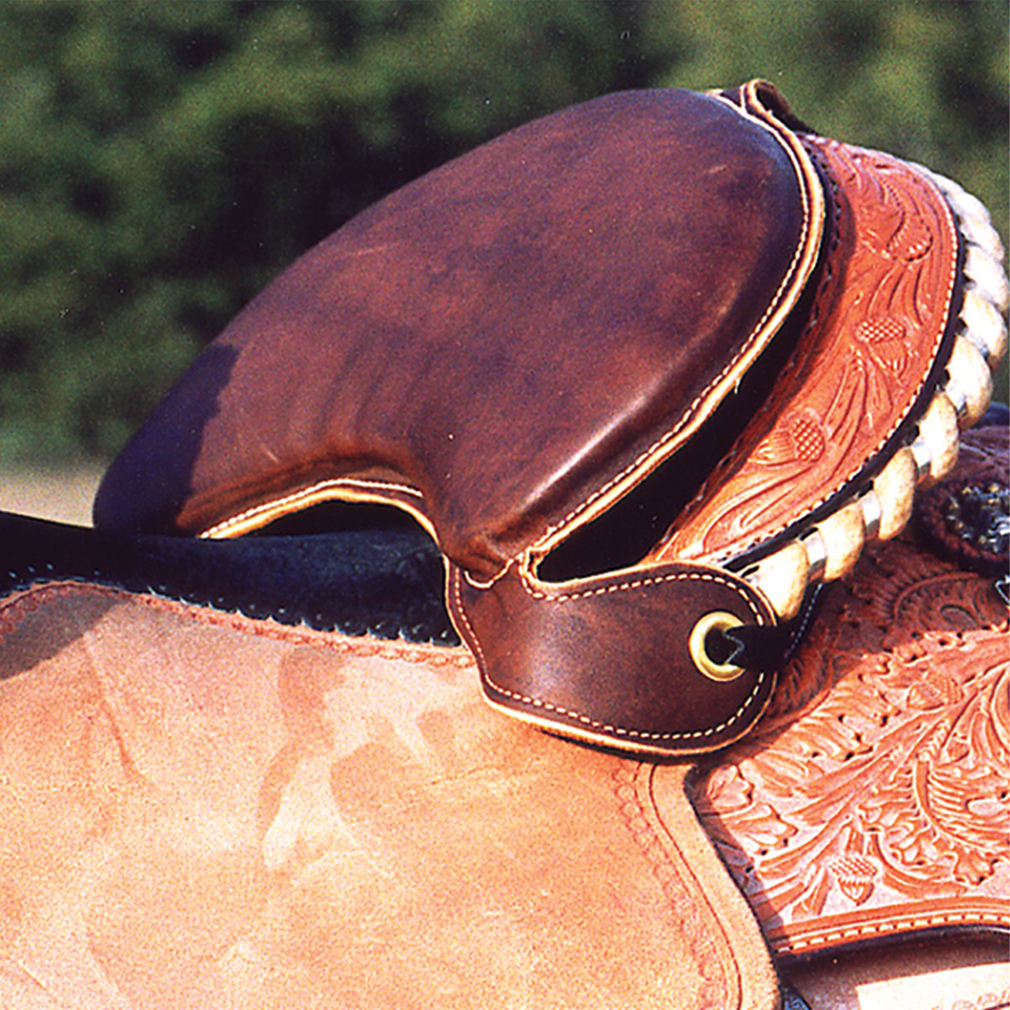 Leather seat shrinker strapped against the cantle of a tooled saddle with a rawhide bound edge. The padded dark leather fills the back of the seat, with trees blurred behind.