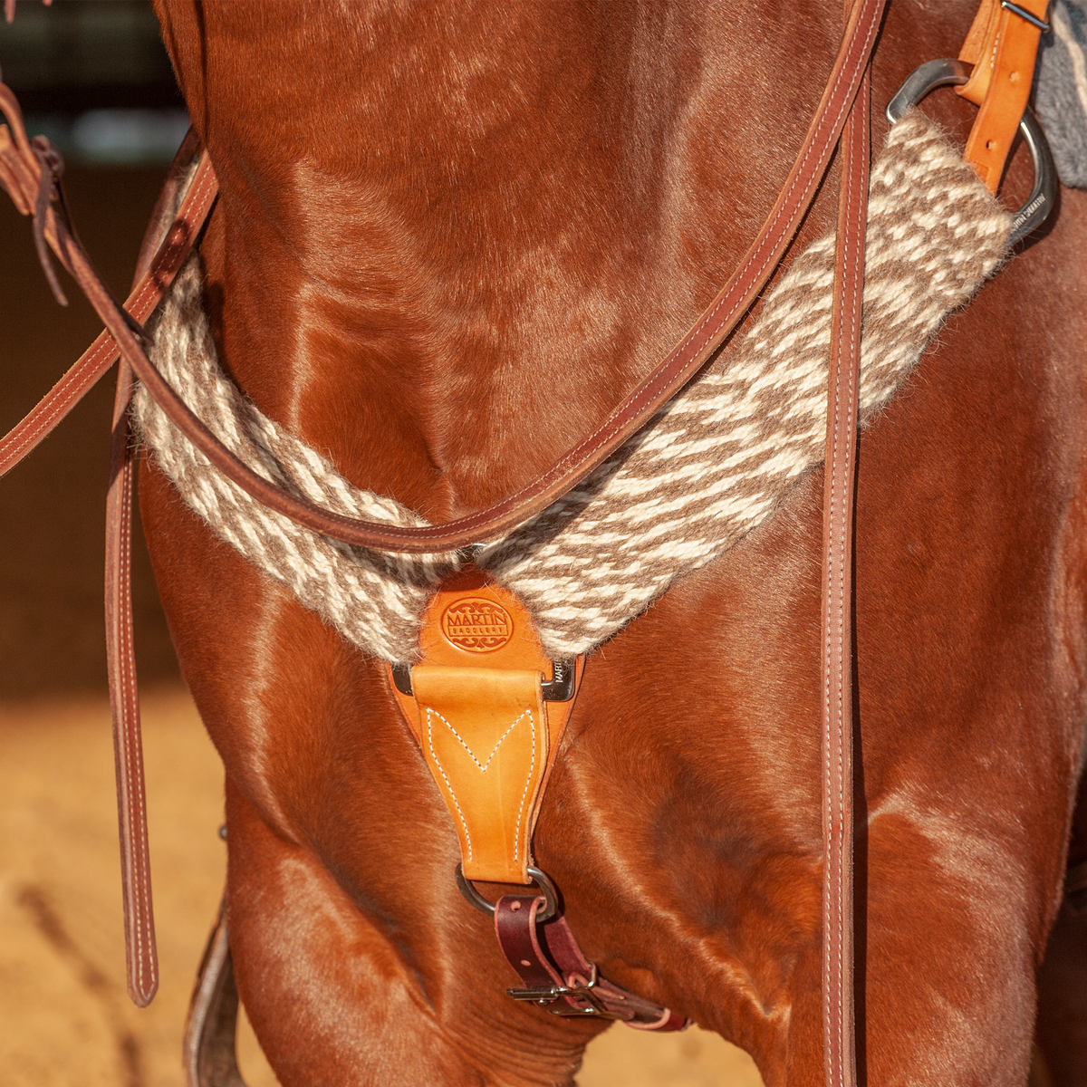 Natural alpaca and mohair breast collar on a horse, seen from the front at the chest. The wide band of natural fiber strands meets a tan leather center piece with a round maker's stamp, with a tie-down strap below.