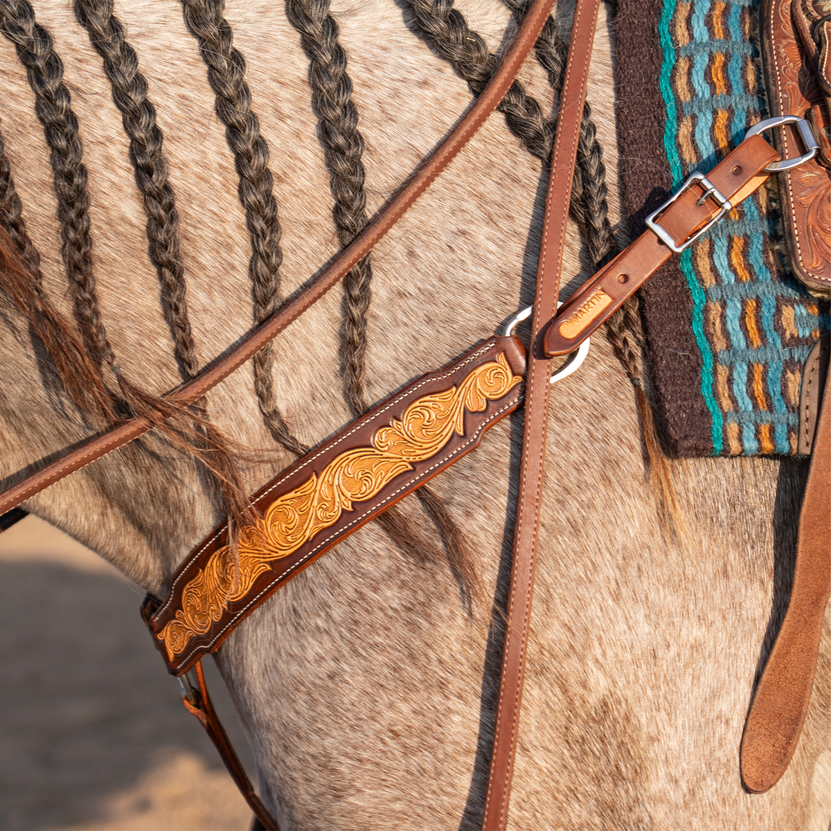Natural breastcollar strap with floral scroll tooling on a gray horse's shoulder, below a braided mane. The chocolate-framed strap connects to a ring and a buckled tug strap at the saddle on this Martin Saddlery breastcollar.
