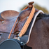Natural roughout rope can strap with chocolate buckstitching hung over the horn of a tooled saddle. The Martin Saddlery strap clips to a black rope can at the horse's shoulder.