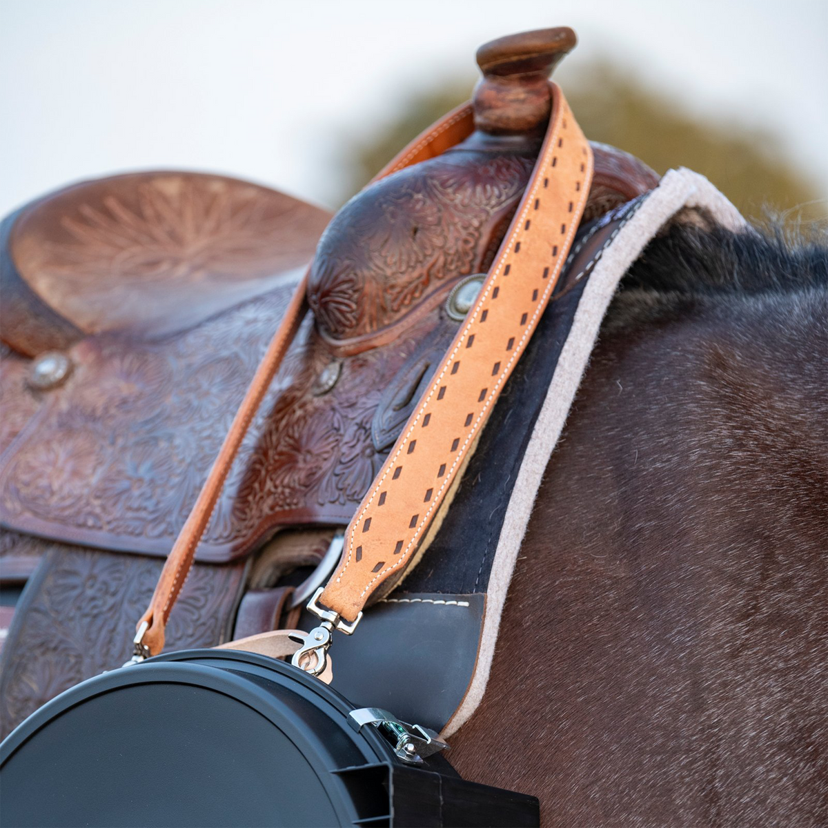 Natural roughout rope can strap with chocolate buckstitching hung over the horn of a tooled saddle. The Martin Saddlery strap clips to a black rope can at the horse's shoulder.