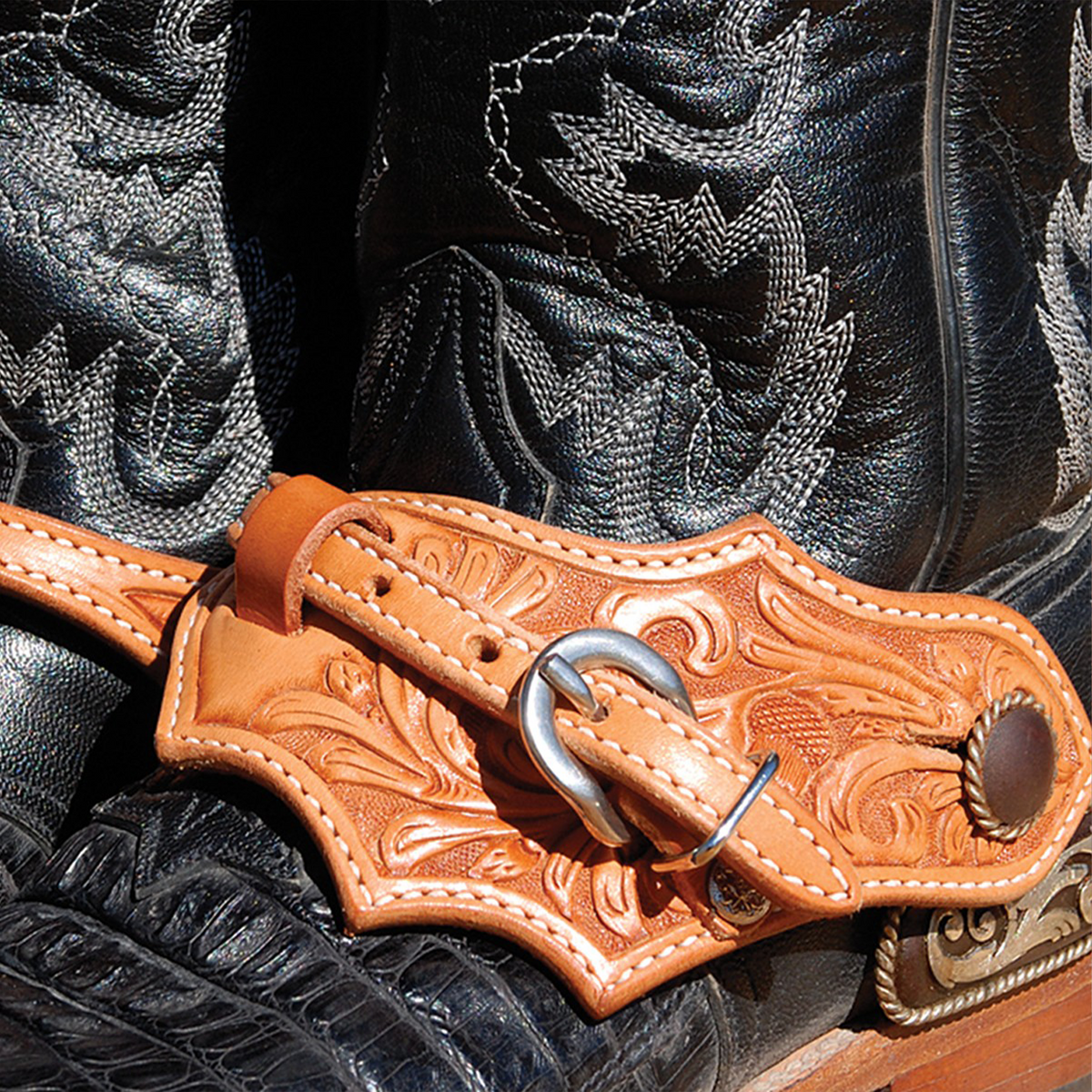Natural oak leaf spur strap with acorn oak tooling buckled over a black cowboy boot, seen up close. White stitching follows the leaf shaped edge around the tooled leather.