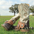 Natural roughout buckaroo spur straps on a pair of spurs worn over cowboy boots standing in grass. The tooled straps buckle across the boot insteps with oak trees behind.