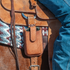 Natural roughout smartphone holder from Martin Saddlery hanging from a saddle beside a rider's leg. A phone sits inside the stitched pouch, which rests against a woven saddle blanket.